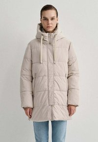 Cappotto imbottito beige con cappuccio, caratterizzato da motivi a trapunta verticali e orizzontali, chiusura anteriore con zip e dettagli con cordino.