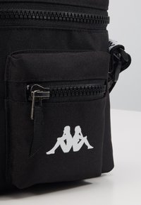 Svart tygpåse med framficka med dragkedja och en vit logotyp som visar två sittande silhuetter ryggen mot ryggen, placerad på en vit yta.