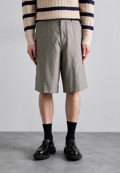 Shorts à carreaux gris avec poches, longueur au-dessus du genou. Portés avec des chaussettes noires et des chaussures noires brillantes. Pull en tricot beige avec des rayures marines.