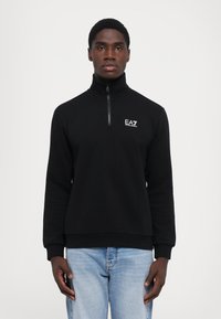 Hanorac negru cu zip, cu guler înalt, din material neted. Prezintă logo-ul EA7 pe piept. Asortat cu blugi de culoare albastru deschis.