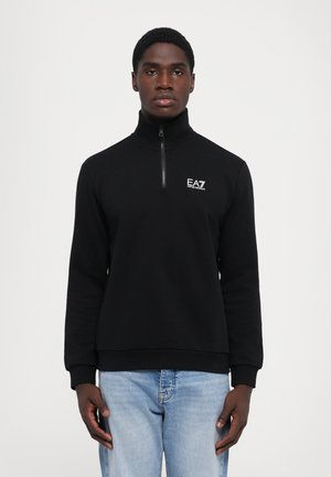 Felpa nera con zip e colletto alto, realizzata in tessuto liscio. Presenta il logo EA7 sul petto. Abbinata a jeans azzurri chiari.