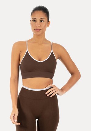 Kvinde stående med hånden på hoften iført brun ribbet sports-BH og leggings med hvid kant på ensfarvet baggrund.