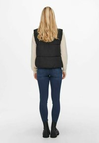 Gilet noir rembourré avec un col montant, porté sur un pull léger, associé à un jean skinny bleu et des bottines noires.