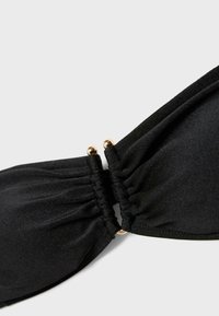 Gros plan sur un haut de bikini en tissu noir avec un fermoir boule doré reliant les deux bonnets triangulaires froncés sur un fond gris clair.