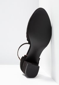 Chaussure noire à petit talon positionnée semelle vers le haut montrant un talon carré texturé et une bride à la cheville sur fond blanc en coin.