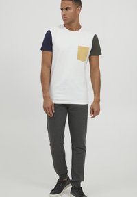 Kortärmad t-shirt med vit kropp, marinblå och olivfärgade ärmar, med en gul bröstficka. Matchas med grå byxor och svarta sneakers.