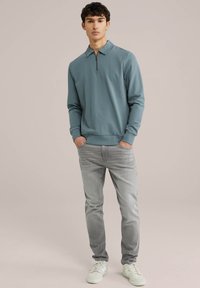 Svijetloplava zip-up majica od mekog materijala, uparena sa svijetlosivim trapericama i bijelim tenisicama. Glatka tekstura i ležeran dizajn.