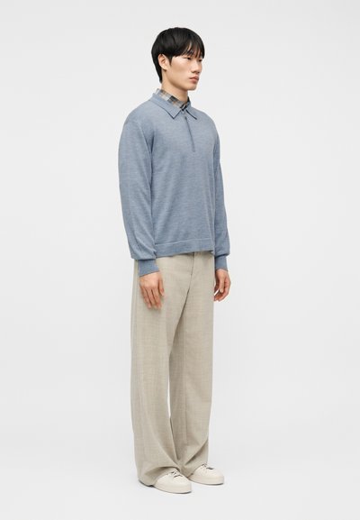 Άνδρας όρθιος, φοράει ανοιχτό μπλε πουλόβερ με φερμουάρ πάνω από καρό πουκάμισο, μπεζ παντελόνι με φαρδιά μπατζάκια και λευκά αθλητικά παπούτσια, με φόντο απλό λευκό.