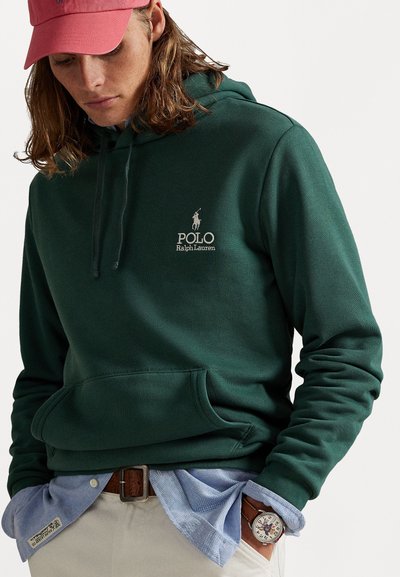 Tmavozelená mikina vyrobená z mäkkého materiálu, s predným vreckom a vyšitým logom. V kombinácii s svetlomodrou košeľou a hnedým opaskom.