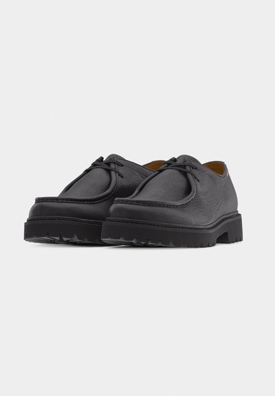 Chaussures à lacets en cuir noir avec surface texturée et semelles en caoutchouc épaisses et côtelées, présentées sous un angle avant-gauche sur fond blanc.