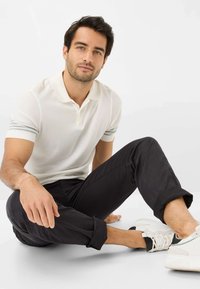 Witte polo met korte mouwen, grijze strepen op de manchetten, gedragen met opgerolde zwarte broek en witte sneaker, zittend tegen een lichte achtergrond.
