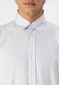 Cămașă button-up cu dungi albastre deschise și albe, cu guler clasic și butoane albe rotunde, având o textură netedă și o croială ajustată.