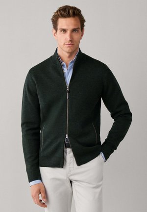 Homme portant un pull zippé vert foncé sur une chemise rayée bleue et un pantalon beige, debout devant un fond clair uni.