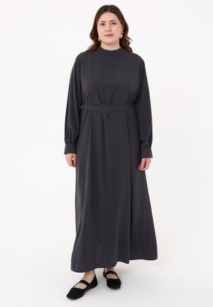 Woman with medium-length brown hair wearing a long dark gray belted dress and black flat shoes, standing against a white background.