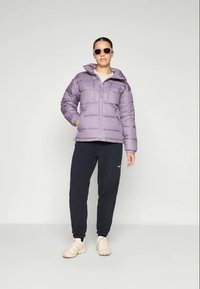 Lys lilla polstret jakke med høy krage, glidelåslukking og sydd mønster, kombinert med svarte joggebukser og beige sneakers.