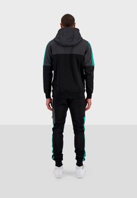 Sudadera negra con un panel superior gris y acentos en azul verdoso, combinada con joggers negros que tienen rayas en azul verdoso y un logo en el muslo.