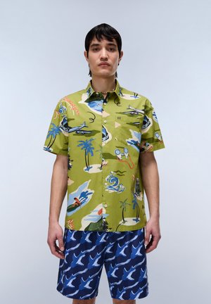 Man met een groen T-shirt met korte mouwen met tropische en vliegtuigprint en blauwe korte broek met een wit vogelpatroon, staand tegen een effen achtergrond.