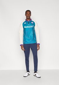 Tirkio spalvos ilgomis rankovėmis sportinis viršus su baltomis rankovėmis, puoštas grafiniu dizainu ir purpuriniais akcentais, derinamas su tamsiai mėlynais sportiniais kelnais ir baltais batais.