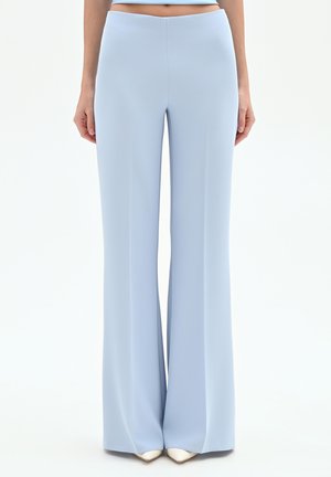 Donna con pantaloni a vita alta e gamba larga di colore azzurro pallido abbinati a scarpe bianche a punta, in piedi contro uno sfondo bianco semplice.