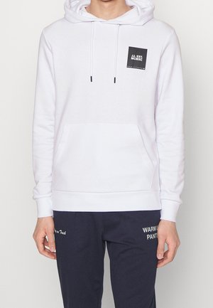 Personne portant un sweat à capuche blanc avec un logo carré noir et un pantalon bleu marine avec un texte blanc sur la cuisse gauche.