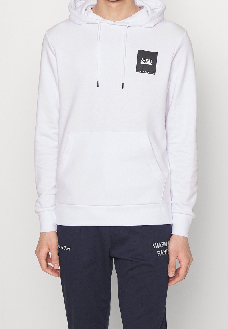 Personne portant un sweat à capuche blanc avec un logo carré noir et un pantalon bleu marine avec un texte blanc sur la cuisse gauche.