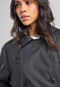 Black waterproof coat with a double-breasted design, featuring a hood, textured fabric, and black buttons, detailed with adjustable drawstrings.