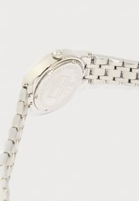 Ασημένιο μεταλλικό ρολόι χειρός με συνδεδεμένο λουράκι, εμφανιζόμενο από πίσω, που δείχνει το χαραγμένο λογότυπο Tommy Hilfiger και τις λεπτομέρειες αντοχής στο νερό.