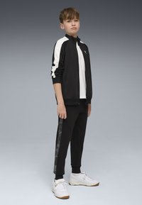 Chaqueta atlética negra con rayas blancas, combinada con pantalones de chándal negros que tienen un panel lateral gris con patrón y zapatillas blancas.