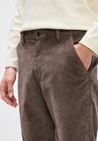 Pantaloni din corduroy maro, cu caneluri verticale, buzunarelaterale și închidere cu nasture. Purtați cu o bluză cu mânecă lungă de culoare deschisă.