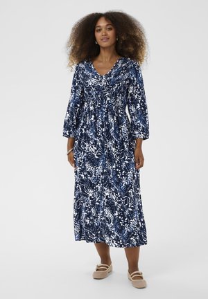 Granatowa sukienka w kwiaty z białymi wzorami, dekolt w serek, rękawy do połowy przedramienia, taliowana, długość do kostek, w zestawieniu z beżowymi butami.