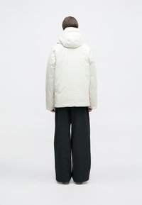 Isik seismas selg ees, seljas heledast beežist kangast kapuutsiga jakk ja laiad mustad püksid, taustal ühtlane valge taust.