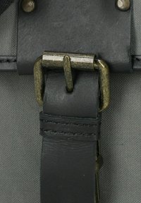 Correa de cuero negro con herrajes de latón unidos a una bolsa de tela gris. Presenta detalles de costura y un diseño de hebilla circular.