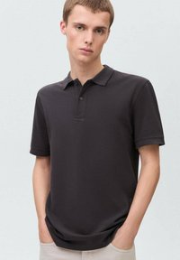 Tummanharmaa lyhythihainen polo-paita, jossa on klassinen kaulus, kaksinappinen placket ja pehmeä, sileä kangas. Kädet taskuissa.