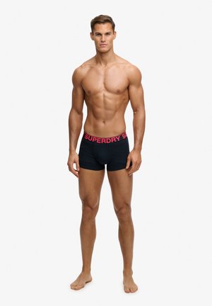 Umięśniony mężczyzna bez koszulki, stojący boso, ubrany w czarne bokserki z czerwoną gumą "SUPERDRY" na białym tle.