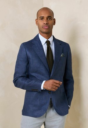 Mann in blauem Blazer, weißem Hemd, dunkler strukturierter Krawatte und hellgrauer Hose steht vor einer beigen Wand und richtet seine Jacke.