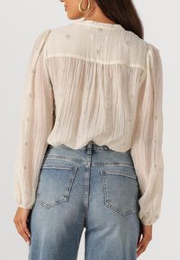Vrouw in een doorschijnende crèmekleurige blouse met parelachtige cirkelbroderie, gecombineerd met hoogzitter blauwe jeans, getoond van de achterkant.