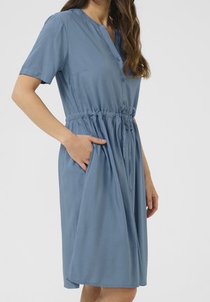Femme portant une robe bleue à manches courtes, boutonnée, longueur genou avec une taille à cordon et une main dans la poche.