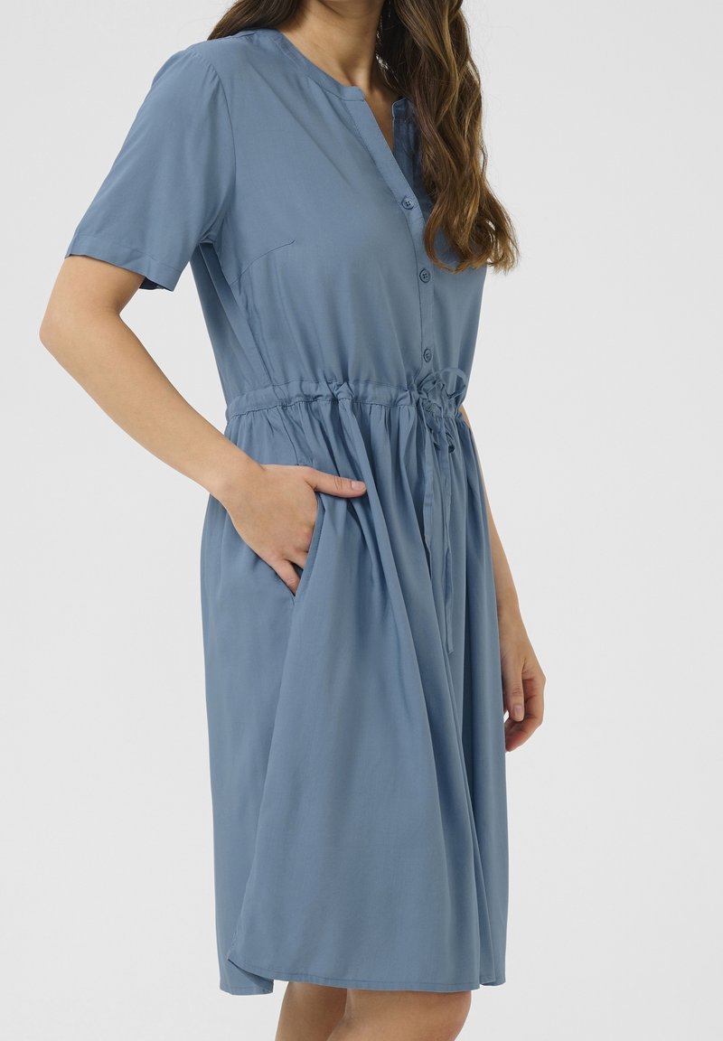 Femme portant une robe bleue à manches courtes, boutonnée, longueur genou avec une taille à cordon et une main dans la poche.