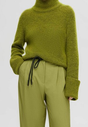 Personne portant un pull col roulé vert et un pantalon taille haute couleur olive clair avec une ceinture à cordon noire, main dans la poche.