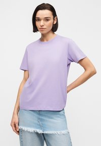 Jonge vrouw met kort donker haar, gekleed in een eenvoudige lavendelkleurige T-shirt en een lichte denimrok met rafelige zoom, staand tegen een witte achtergrond.