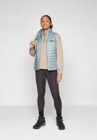 Lyseblå puffervest med høyt krage, båret over en beige zip-up genser, kombinert med svarte leggings og tursko.
