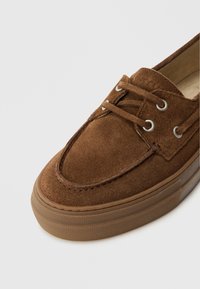 Hnědá semišová bota s kulatou špičkou, šněrováním, kovovými průchodkami a strukturovanou gumovou podrážkou. Viditelné švy a měkký povrch.