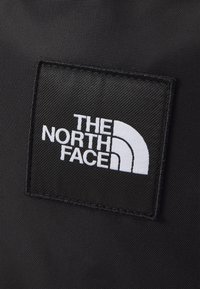 Czarna tkaninowa naszywka z białym napisem "THE NORTH FACE" oraz zakrzywionym designem logo; teksturowane tło. Krawędzie są przeszyte.