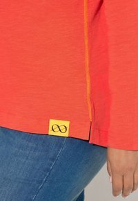 Rotes Langarmshirt mit gelben Nähten und einem gelben Etikett mit Unendlichkeitssymbol. Das Material wirkt weich und strukturiert.