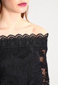 Top negro de encaje con hombros al descubierto que presenta intrincados patrones florales, bordes en forma de ondas y una tela texturizada.