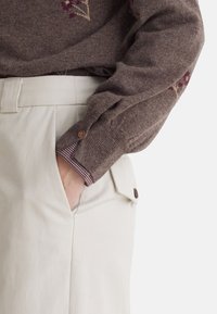 Maglione marrone lavorato a maglia con ricami floreali, abbinato a pantaloni color crema con tasca laterale e dettaglio con bottone sulla vita.