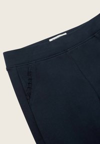 Námornícke modré nohavice s hladkým povrchom, s elastickým pásom a volánovými detailmi na bočných vreckách. Štítok viditeľný na vrchu.