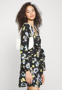 Vestido floral de mangas longas em preto, com flores amarelas e lavanda; uma bolsa branca com alça de corrente repousa no ombro.