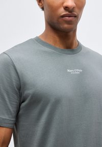 Άνδρας που φοράει γκρι μπλουζάκι με στρογγυλή λαιμόκοψη με την επιγραφή "Marc O’Polo EST. IN STOCKHOLM" στο στήθος, εμφανιζόμενος από τον λαιμό μέχρι το μέσο του κορμού.