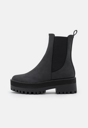 Bottines Chelsea noires en matériau lisse, dotées de panneaux latéraux élastiques, d'une semelle épaisse à texture rugueuse et d'une languette à l'arrière.